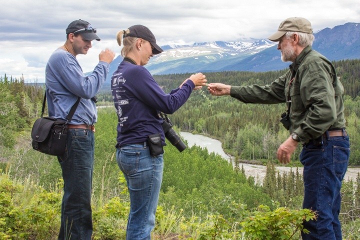 Three people exchange items with binoculars outdoors, overlooking a scenic forest and river valley.