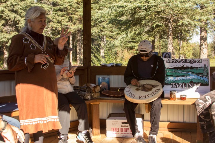 People on a wooden porch, one holding a drum, surrounded by nature and Native Village signage.