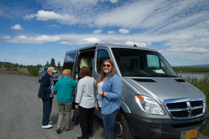 People boarding a gray van on a scenic rural road.
