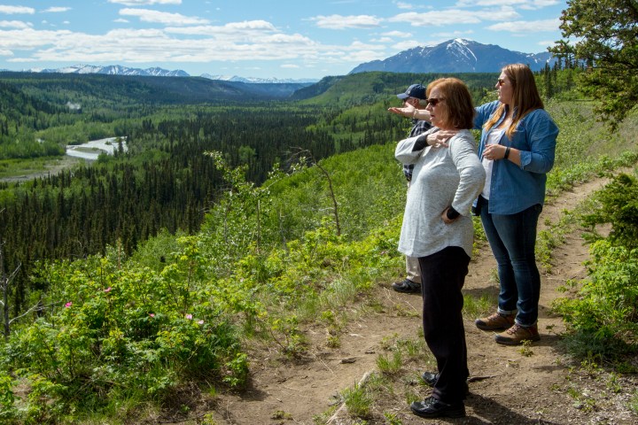 Three people admire a scenic mountain view with a river below.