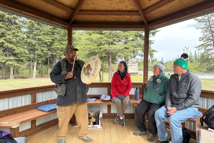Person holds a drum in a wooden gazebo with three seated people listening.