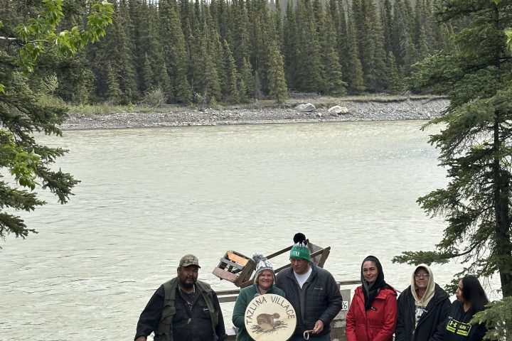 Five people by a river, one holding a drum, surrounded by pine trees.