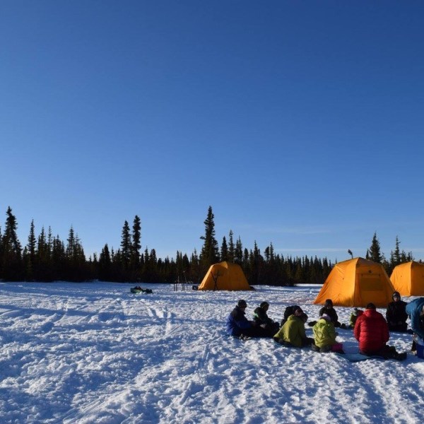People seated near orange tents on a snowy landscape with trees.