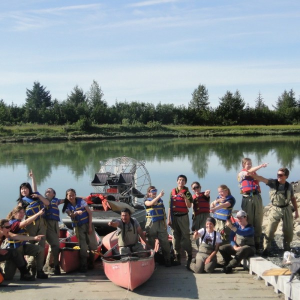 Group in life vests pose with paddles by a river, next to a red canoe and an airboat.