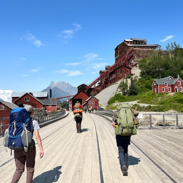 Hikers with backpacks on a wooden path towards red buildings on a hillside under a clear blue sky.